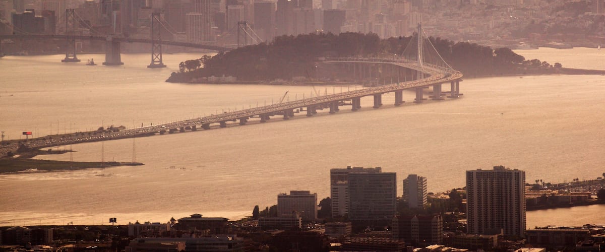 A cityscape with a prominent bridge spanning a river, surrounded by high-rise buildings and a hilly backdrop.
