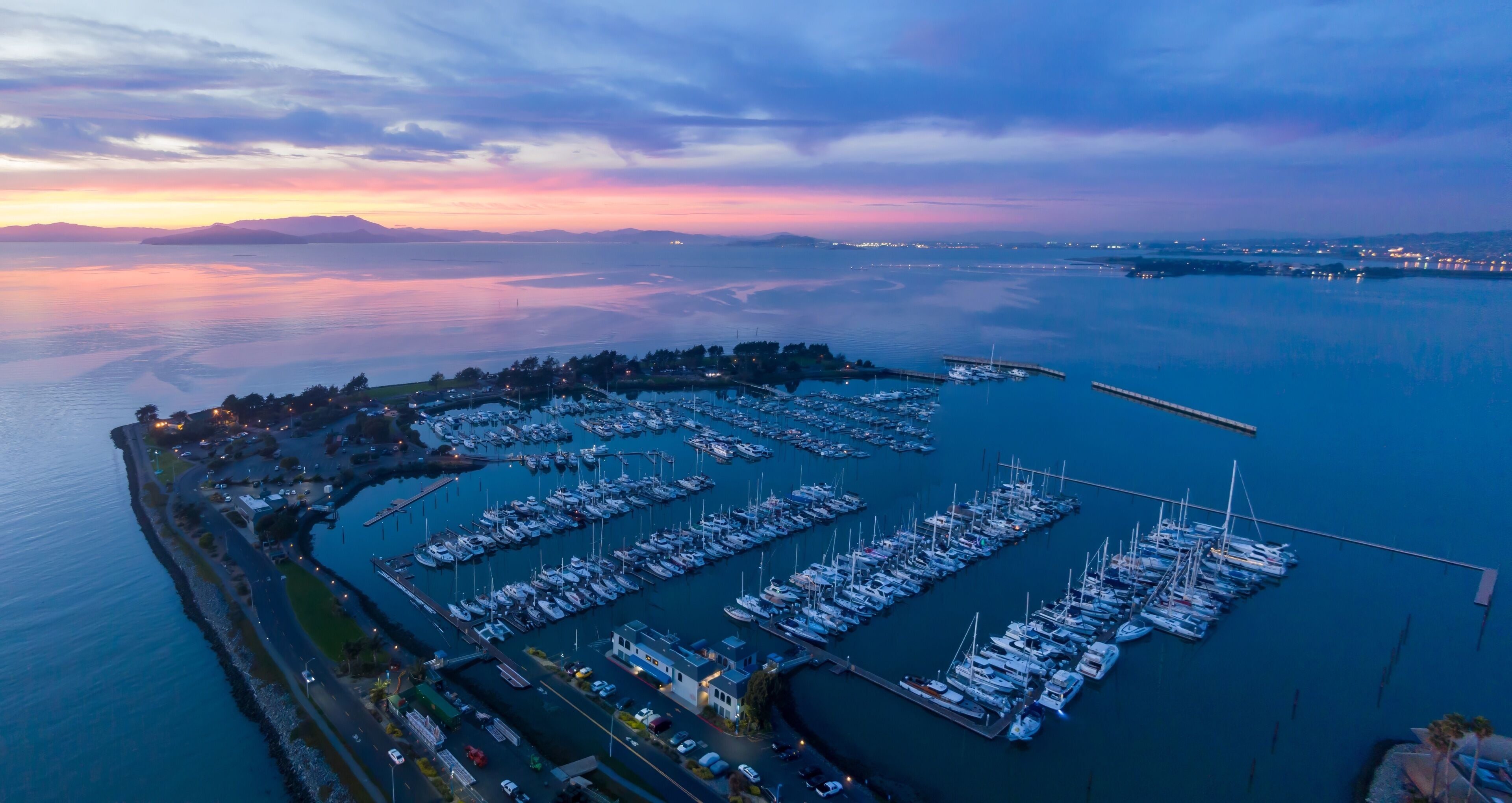 A marina with numerous boats docked, surrounded by calm waters and a distant sunset.