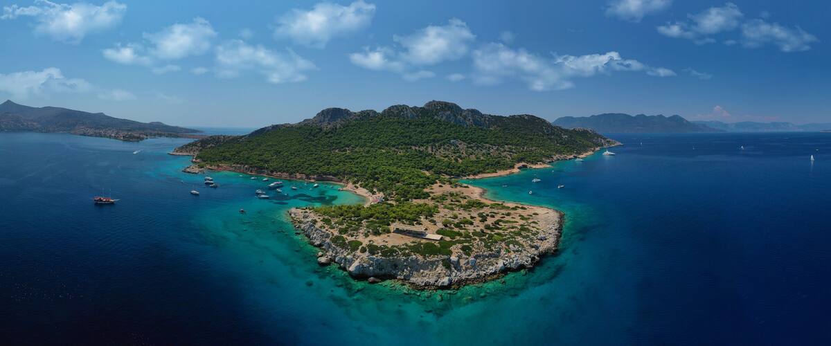 An island surrounded by clear blue waters with boats, a rocky coastline, and lush greenery.