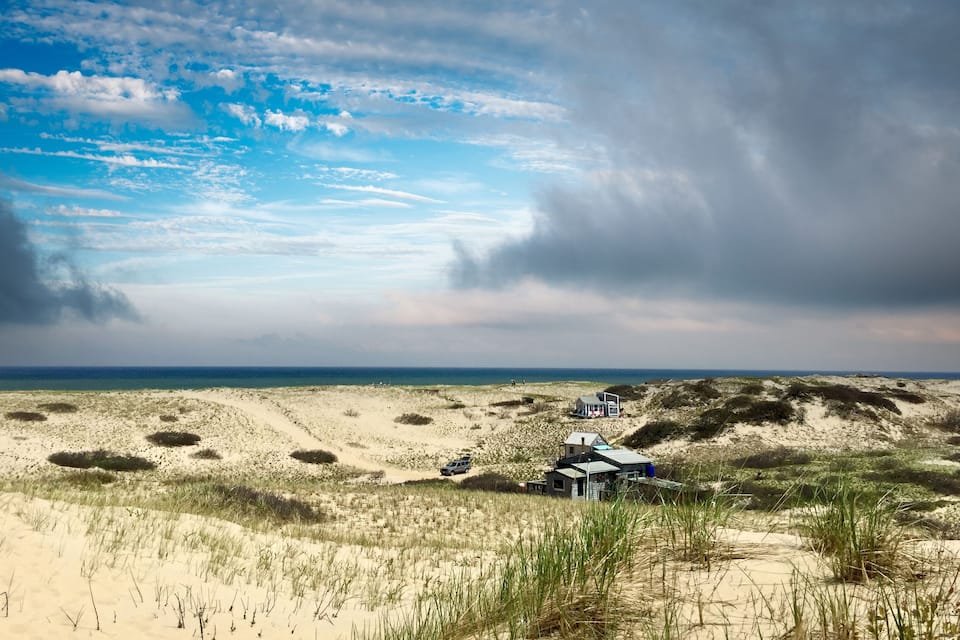 Un paysage côtier avec des dunes de sable, une petite maison et un ciel dégagé.