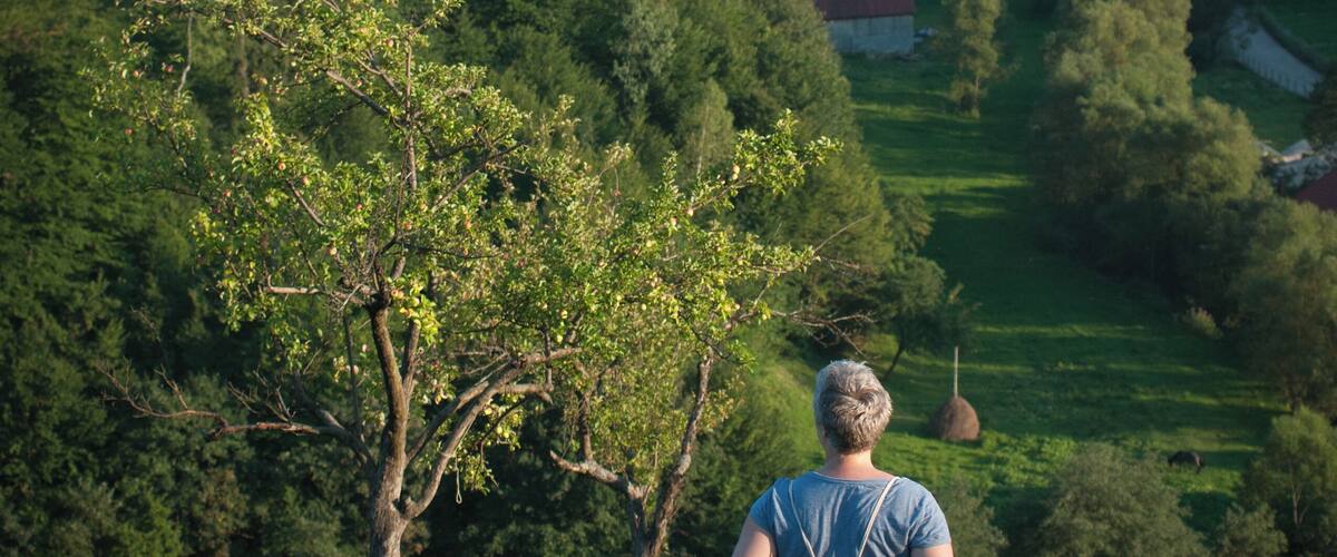 A woman standing on a grassy hill overlooking a valley with trees and a red-roofed building.