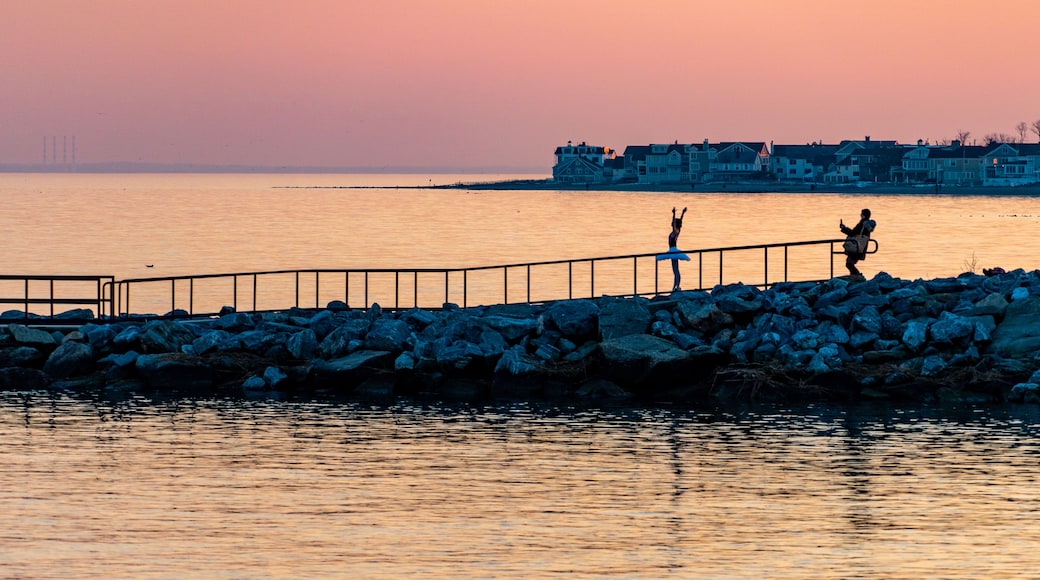 A sunset over a calm body of water with a rocky pier and buildings in the background.