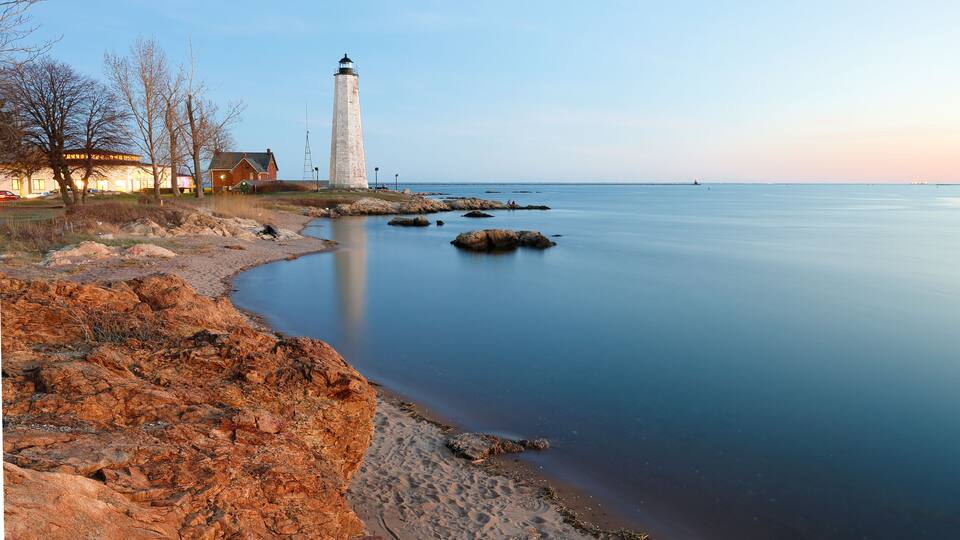 A coastal landscape with a lighthouse, rocky shore, calm water, and a building with lights.