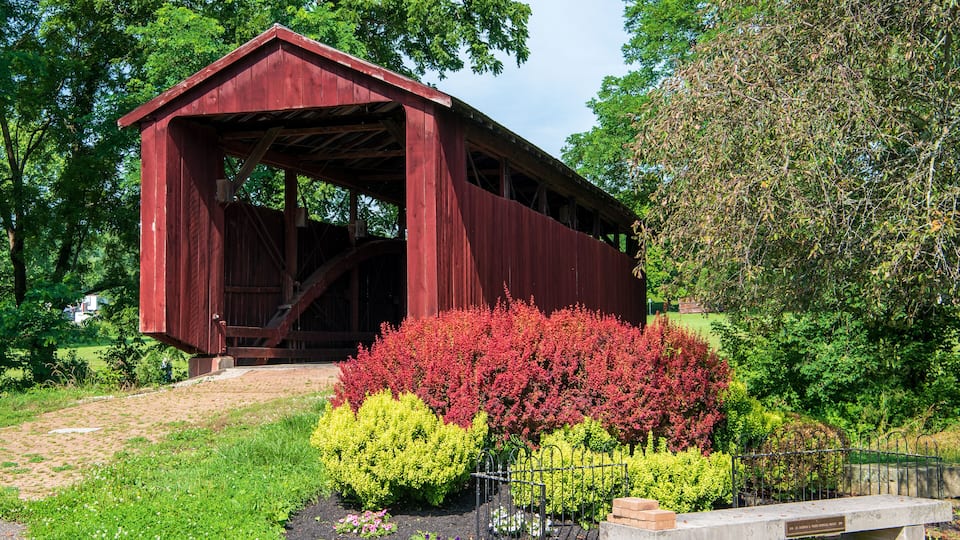 A red covered bridge, surrounded by vibrant red and yellow flowering bushes, set in a lush green landscape.