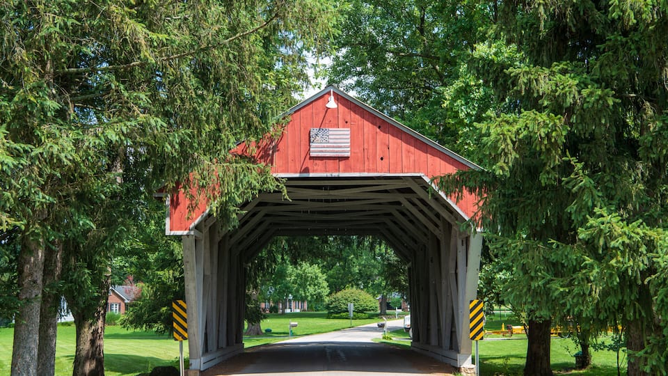 A red covered bridge with a flag, surrounded by trees and a clear road.