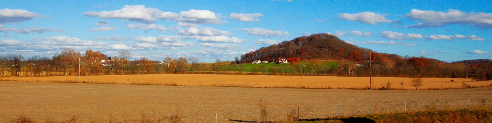 A rural landscape with a hill, open fields, and a clear blue sky with scattered clouds.