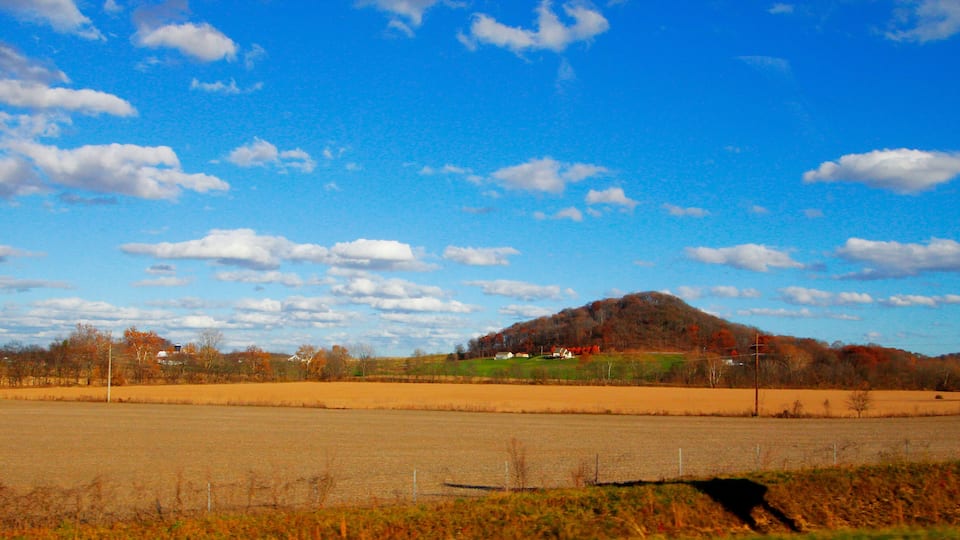 A rural landscape with a hill, open fields, and a clear blue sky with scattered clouds.