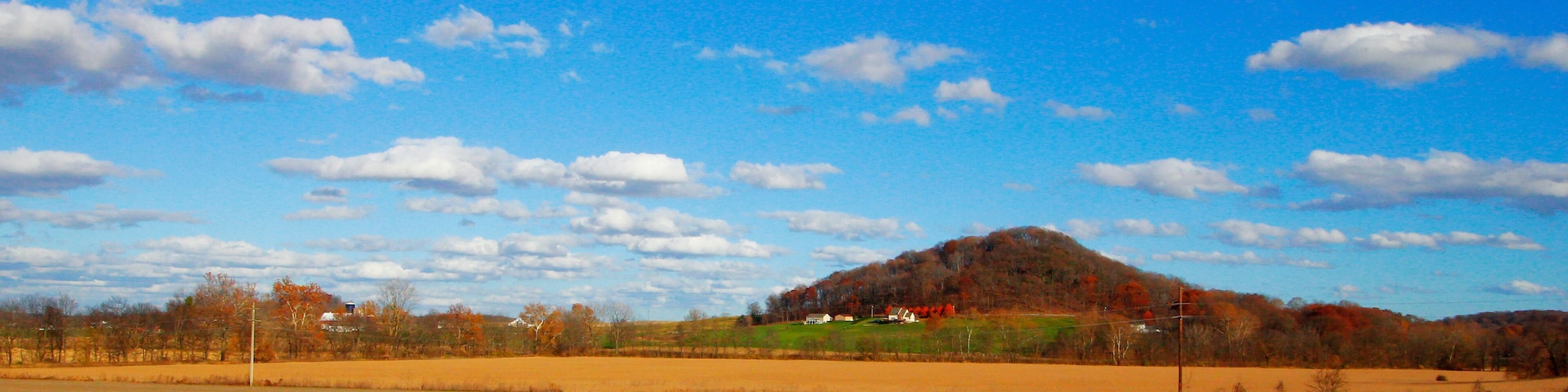 A rural landscape with a hill, open fields, and a clear blue sky with scattered clouds.