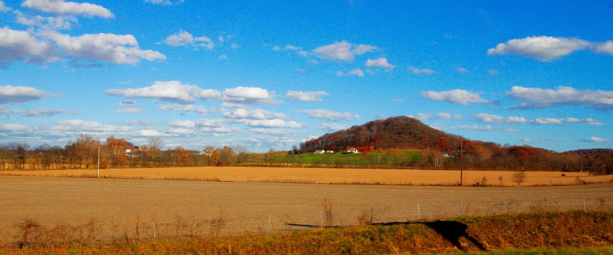 A rural landscape with a hill, open fields, and a clear blue sky with scattered clouds.