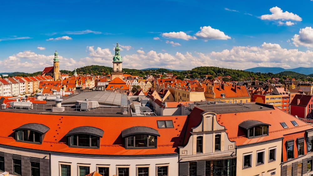 Eine Stadtansicht mit roten Dächern, einer markanten Kirche mit grünem Kirchturm und einem bergigen Hintergrund unter blauem Himmel.