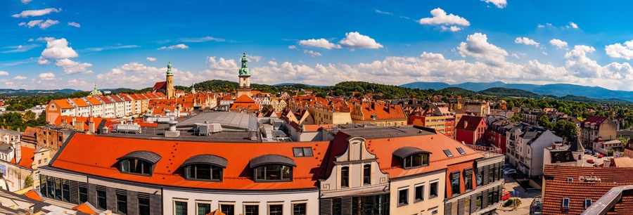 A cityscape with red-roofed buildings, a prominent church with a green spire, and a mountainous backdrop under a clear sky.