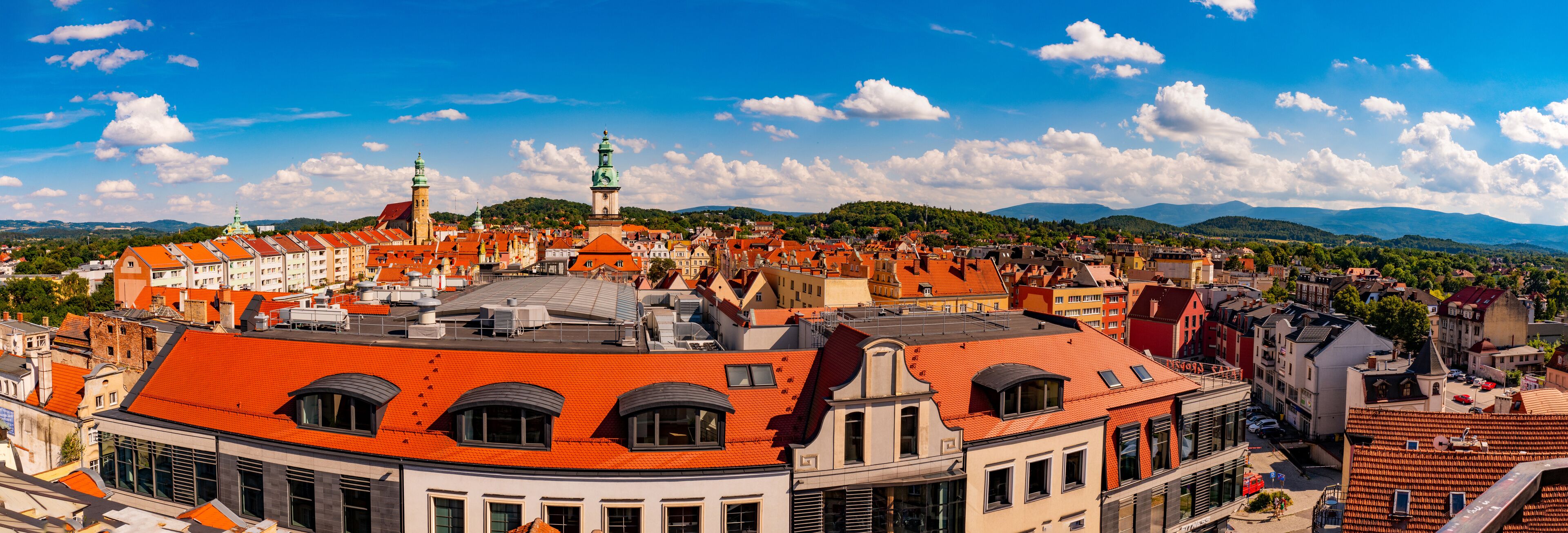 A cityscape with red-roofed buildings, a prominent church with a green spire, and a mountainous backdrop under a clear sky.
