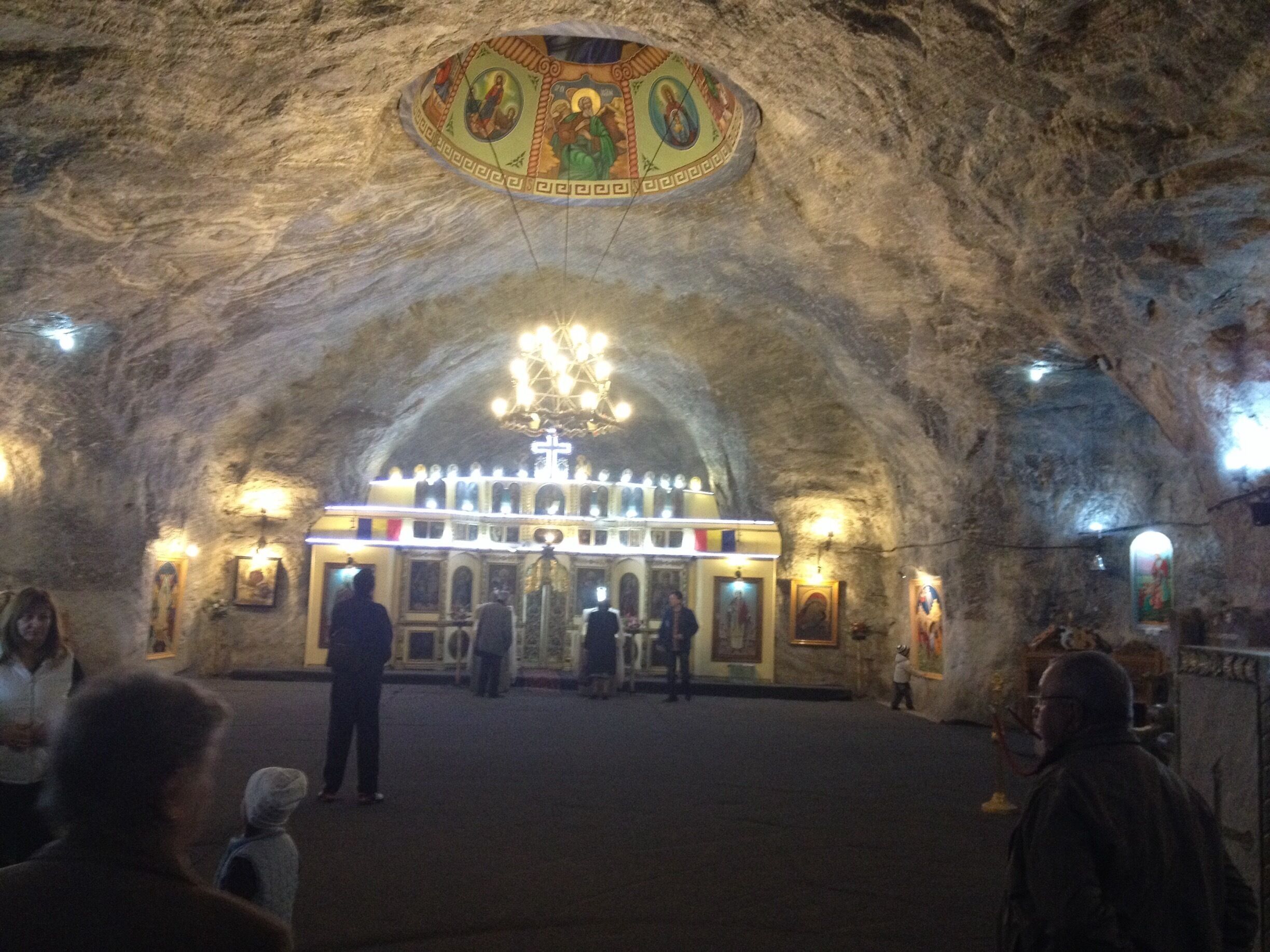 An interior of a cave church with a central altar, chandeliers, and religious paintings.