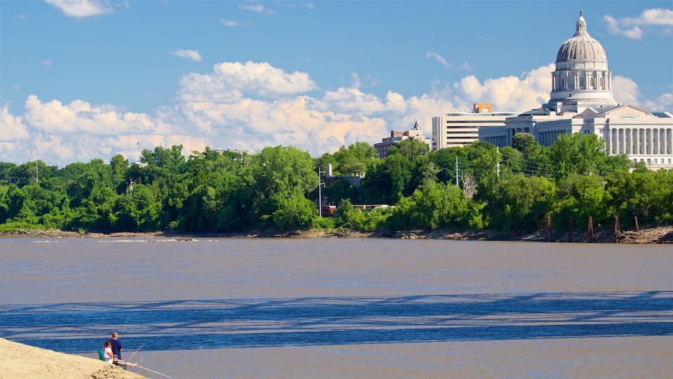 A river with a sandy beach, a person fishing, and a large building with a dome in the background.
