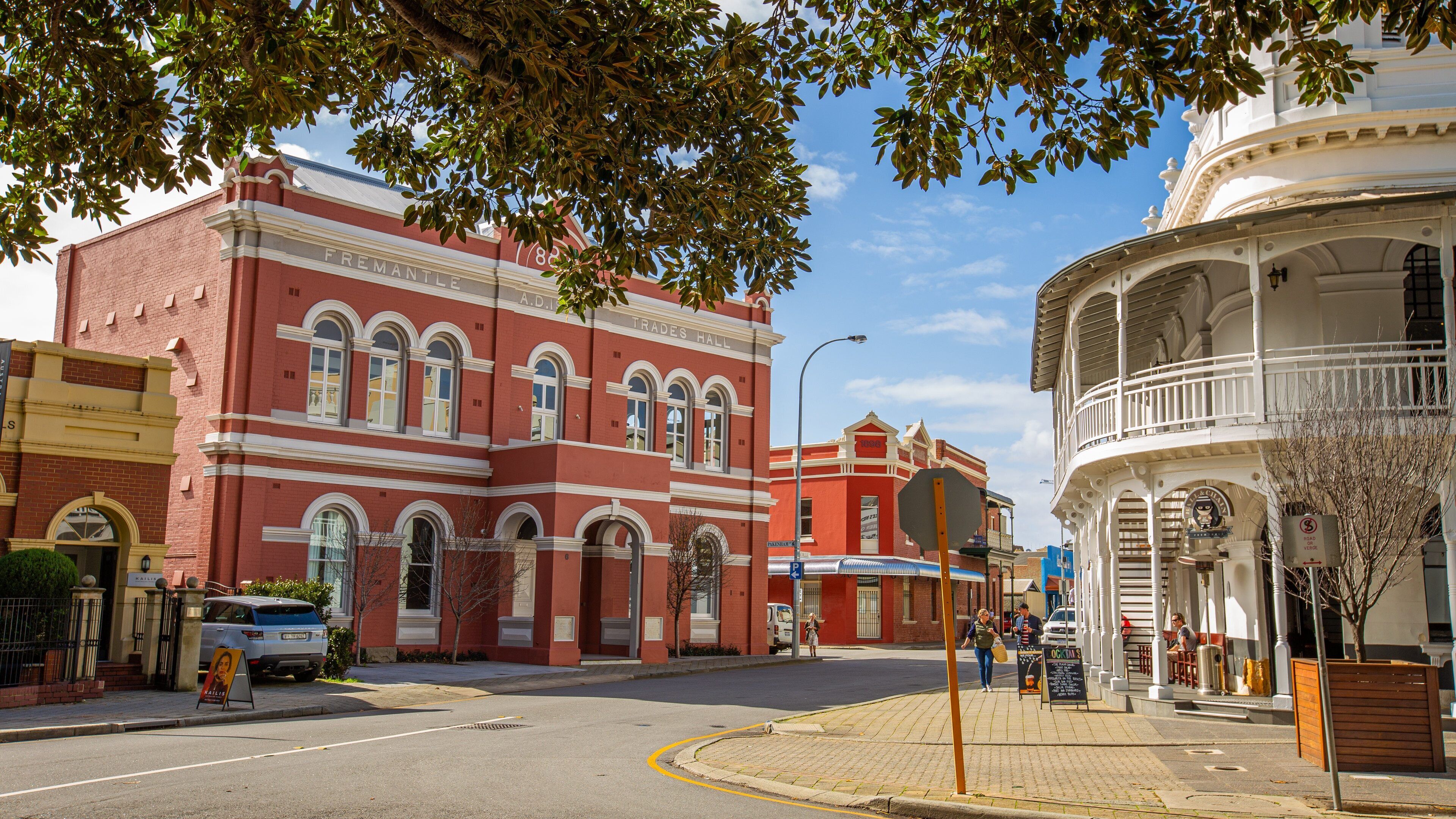 A street with historic buildings, including a red brick building labeled 'Trades Hall' and a white building with a balcony.