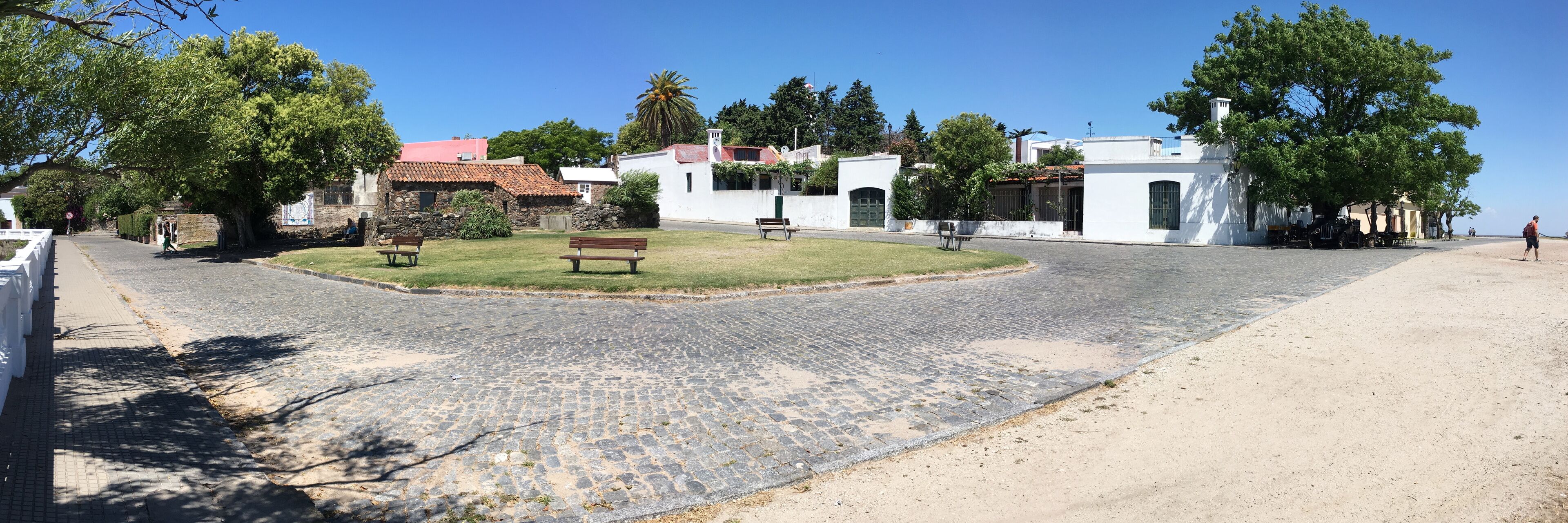 A cobblestone street with benches, white buildings, and trees.