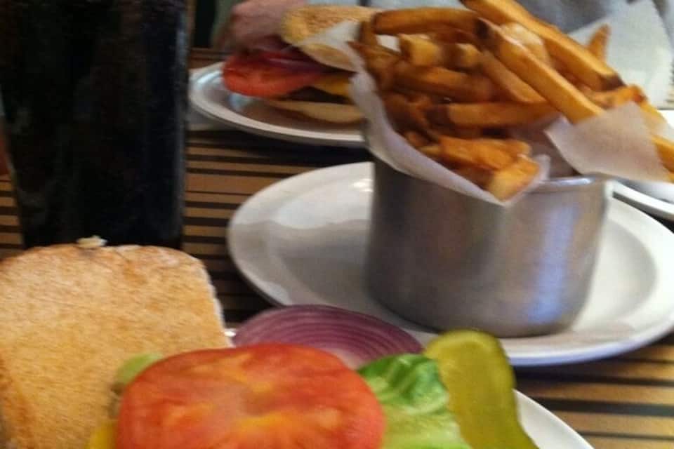 A sandwich with lettuce, tomato, and pickle, a glass of soda, and a basket of fries on a table.
