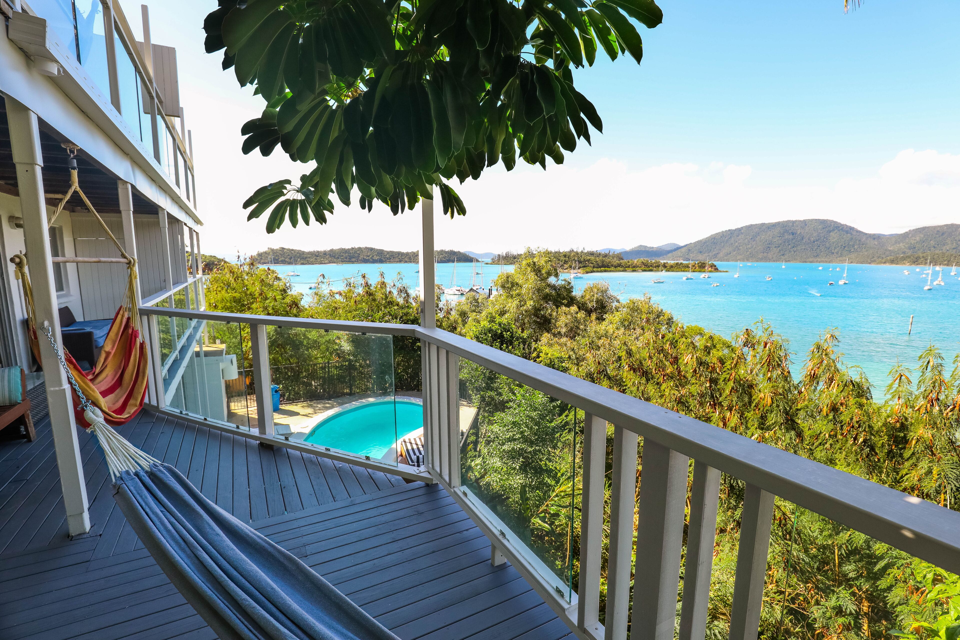 Um deck de madeira com uma rede, com vista para um mar azul e límpido com barcos e uma piscina.