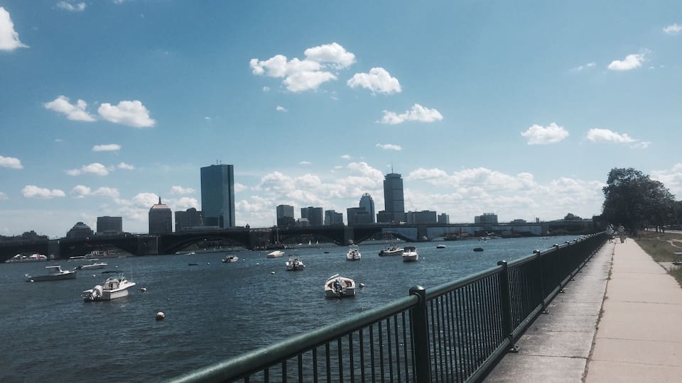 A river with boats, a city skyline, and a pedestrian walkway.