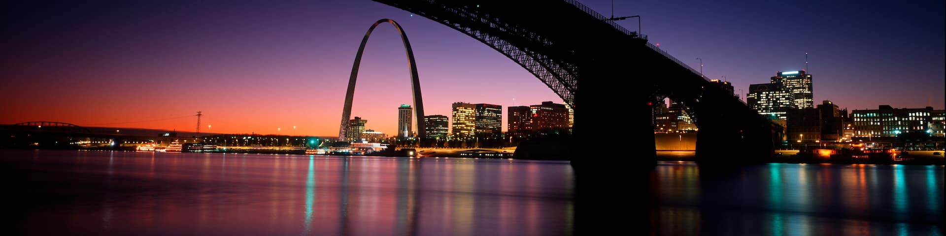 A city skyline at dusk with a prominent bridge arching over a body of water.