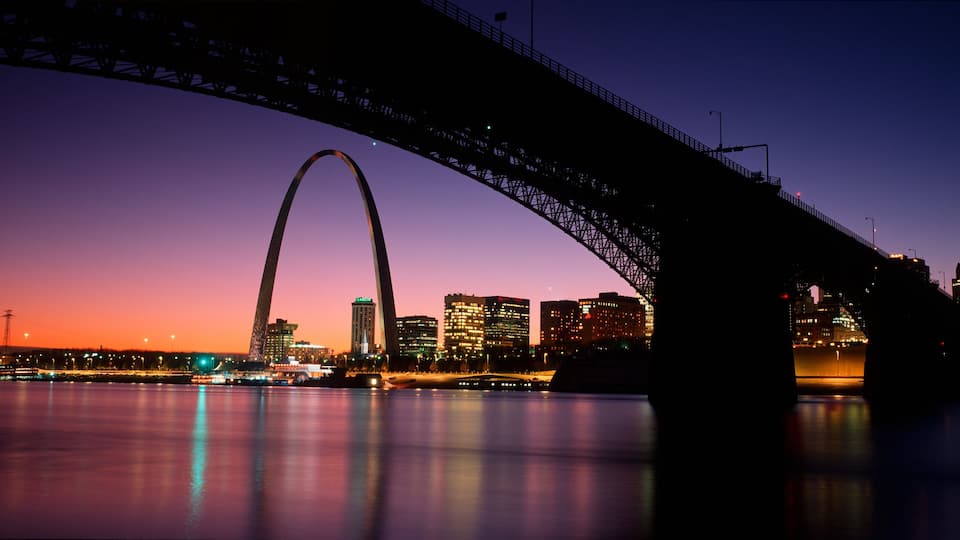 A city skyline at dusk with a prominent bridge arching over a body of water.