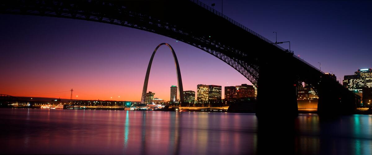 A city skyline at dusk with a prominent bridge arching over a body of water.