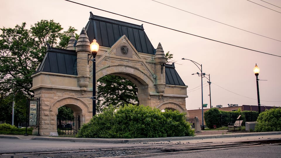 A stone archway with a metal roof, surrounded by greenery and street lamps.