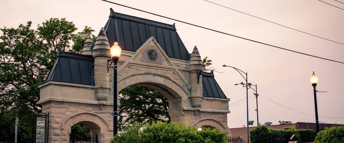 A stone archway with a metal roof, surrounded by greenery and street lamps.