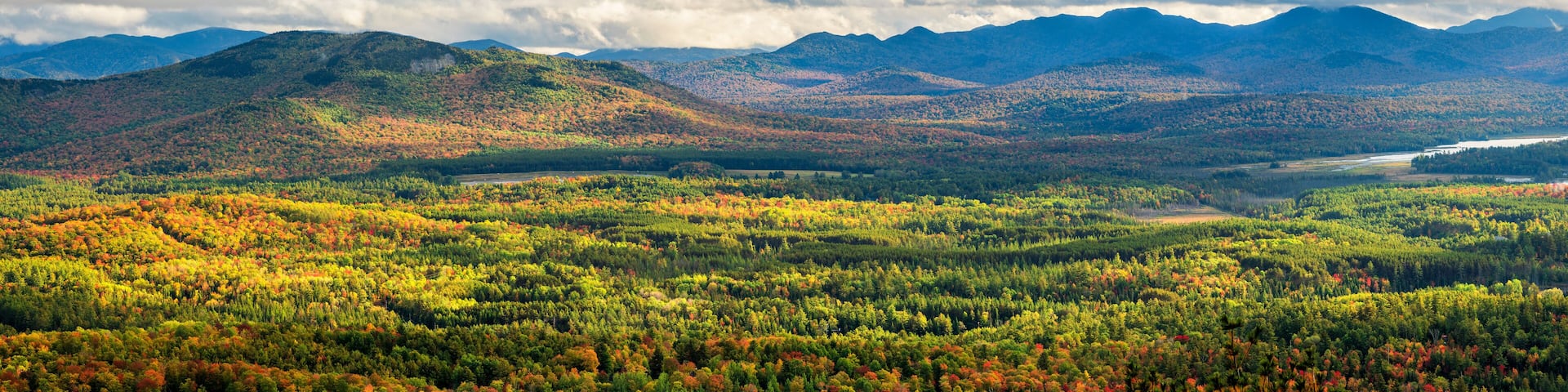 A forested valley with autumn foliage and distant mountains under a cloudy sky.