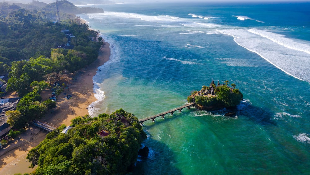 Eine Küstenlandschaft mit Strand, bewaldetem Gebiet und einer Brücke, die zwei Landmassen über dem Ozean verbindet.