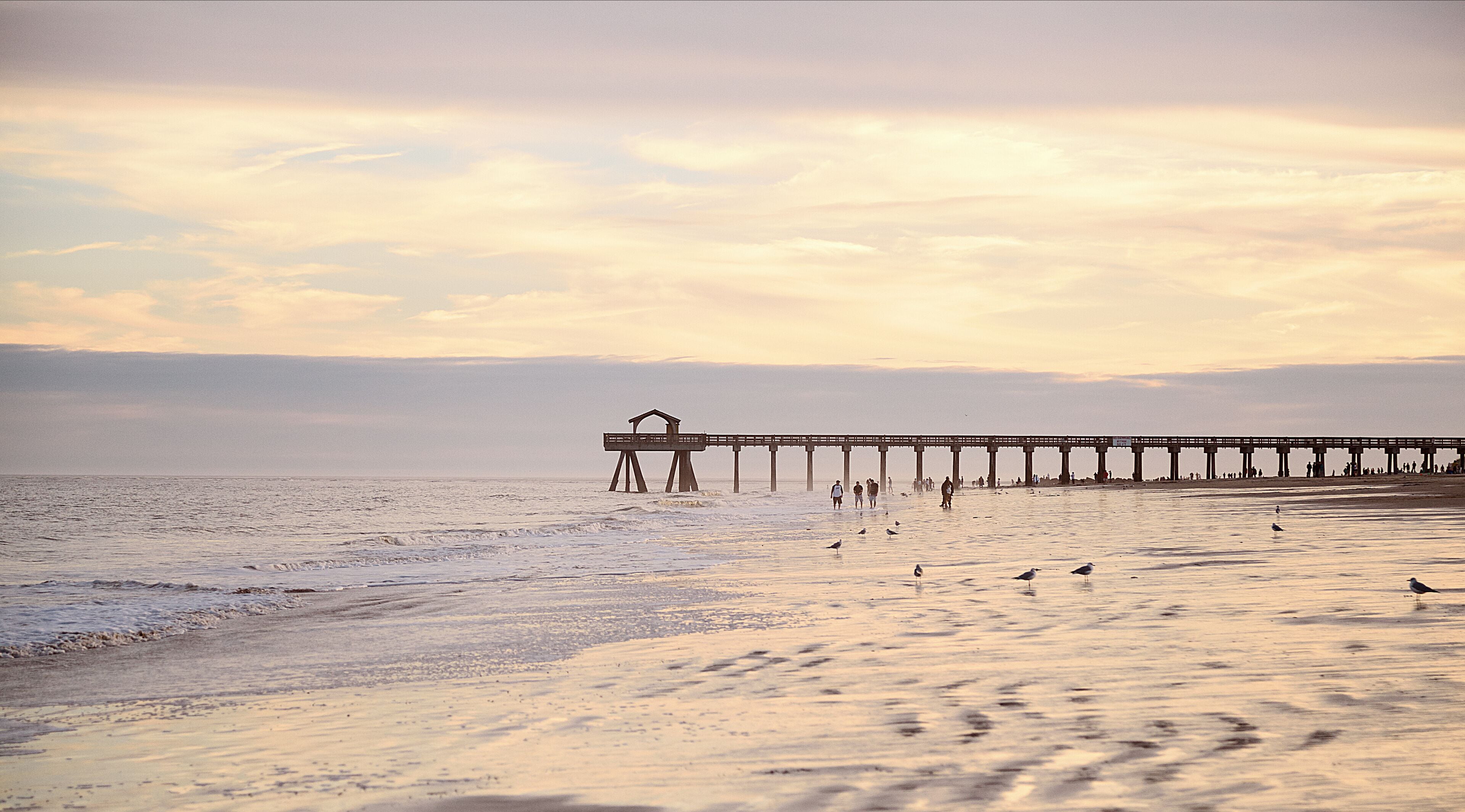 Ocean pier jetty at spectacular sunset. People walking on beach. Peaceful scene, calming waves, pastel cloudy sky, coast. Soft light