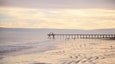 Ocean pier jetty at spectacular sunset. People walking on beach. Peaceful scene, calming waves, pastel cloudy sky, coast. Soft light