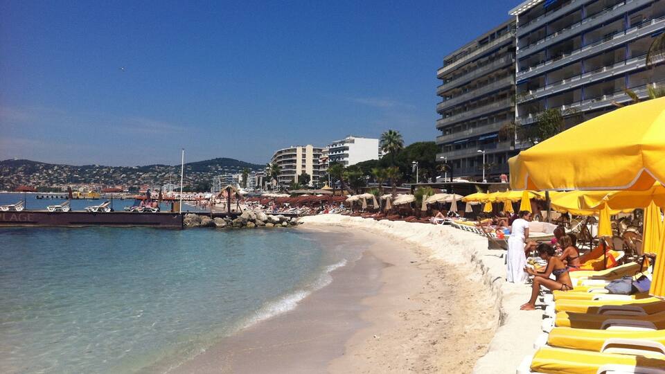 A beach with yellow umbrellas and lounge chairs, a pier, and buildings in the background.