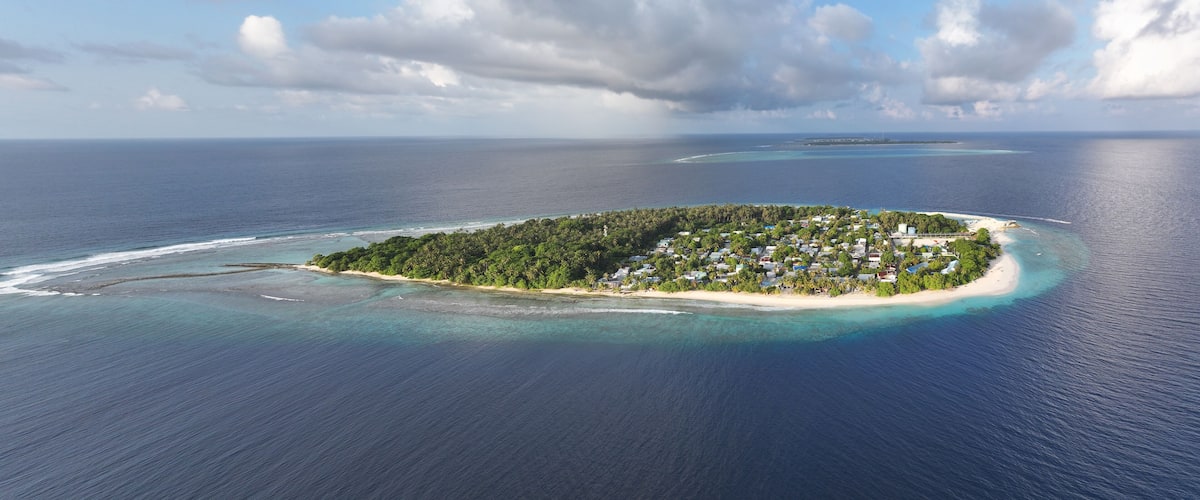 An aerial view of a tropical island with a small settlement, surrounded by clear turquoise waters and a distant coral reef.