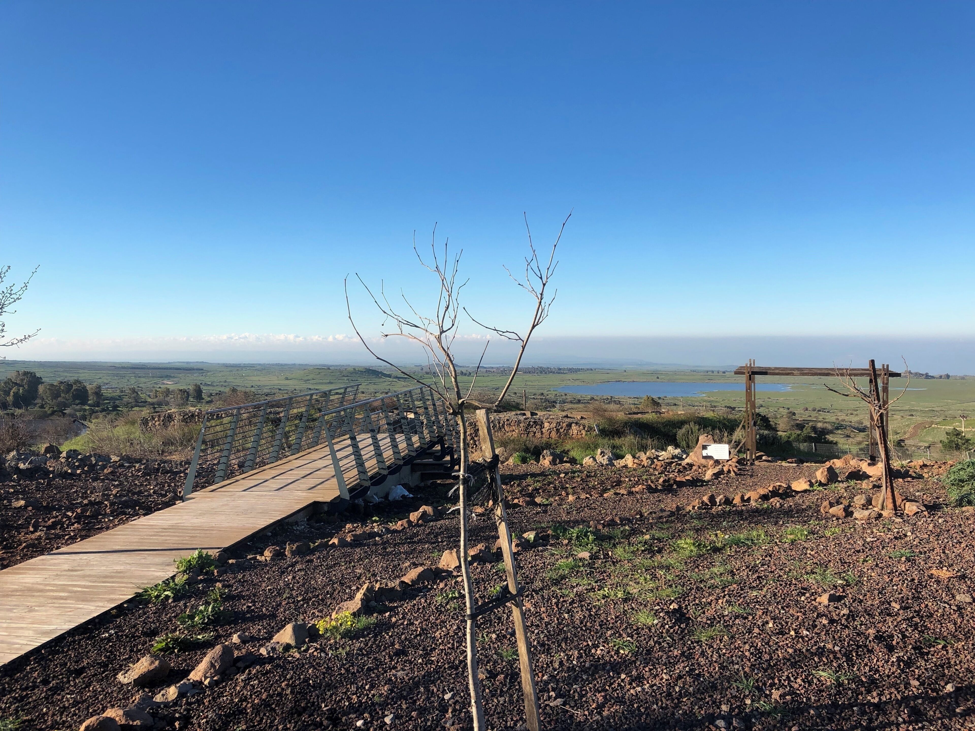 A wooden boardwalk, rocky terrain, and a distant view of a lake and fields under a clear blue sky.