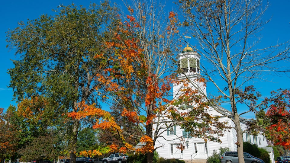 A white church with a gold dome, surrounded by autumn trees with orange leaves, and parked cars in a suburban setting.
