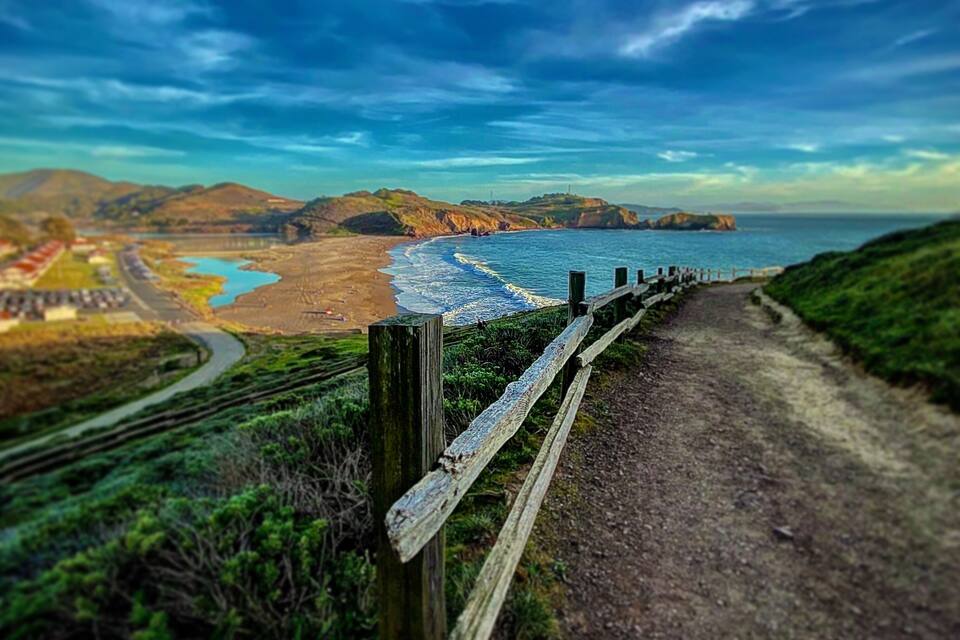 Een kustlandschap met een onverhard pad, houten omheining en een strand.