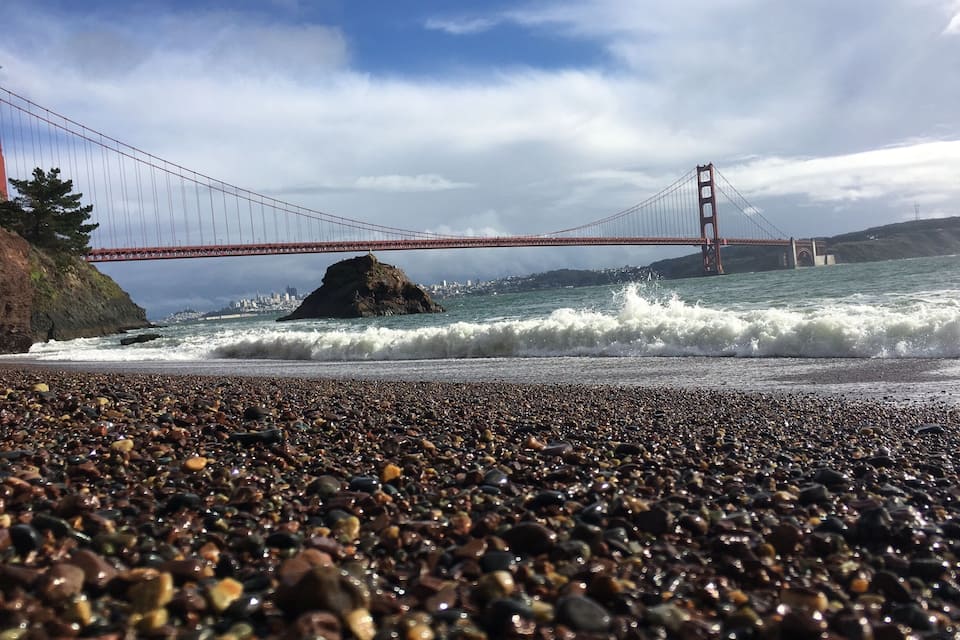 Una playa de guijarros con olas rompiendo en la orilla, un promontorio rocoso y el Puente Golden Gate al fondo.