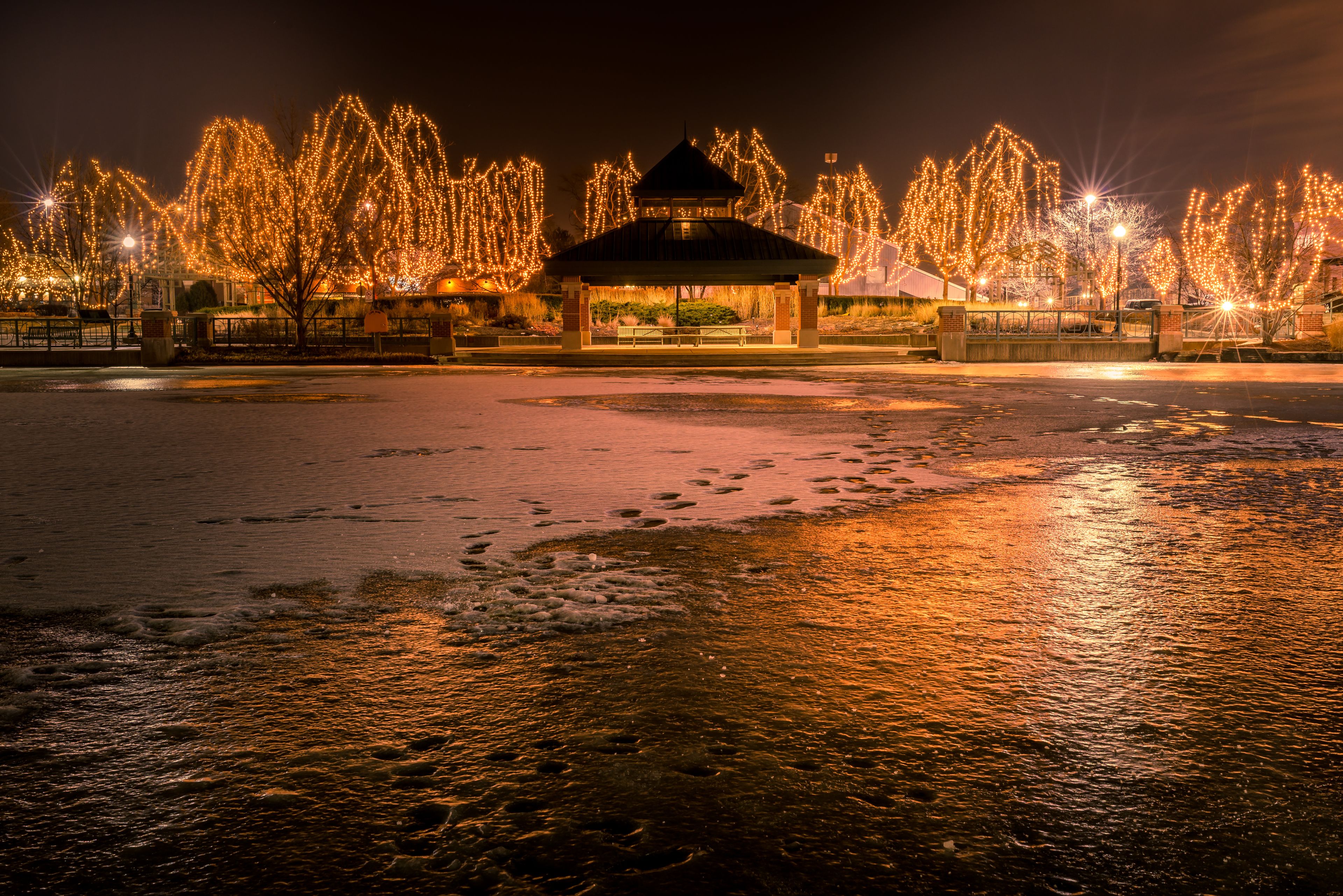 A snow-covered beach at night with a gazebo and trees decorated with lights.