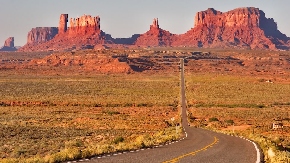 A winding road in a desert landscape with red rock formations in the background.