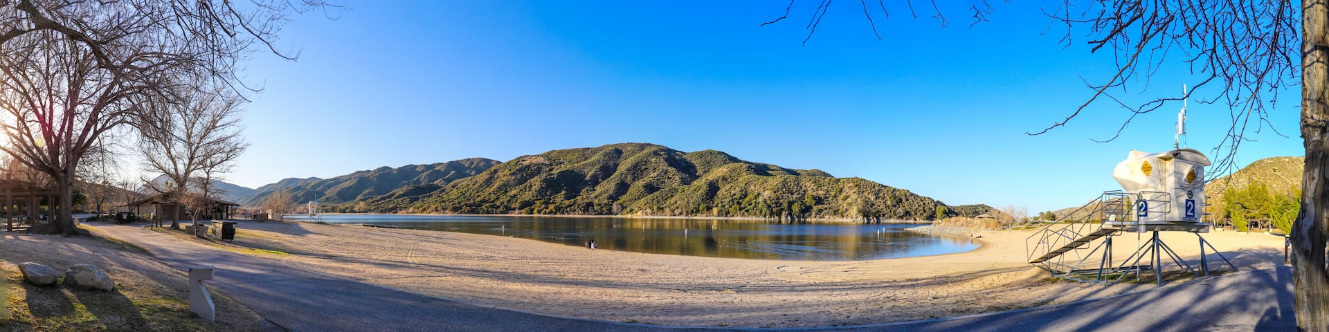 A lakeside road with a sandy beach, a forested hill, and a lifeguard tower.