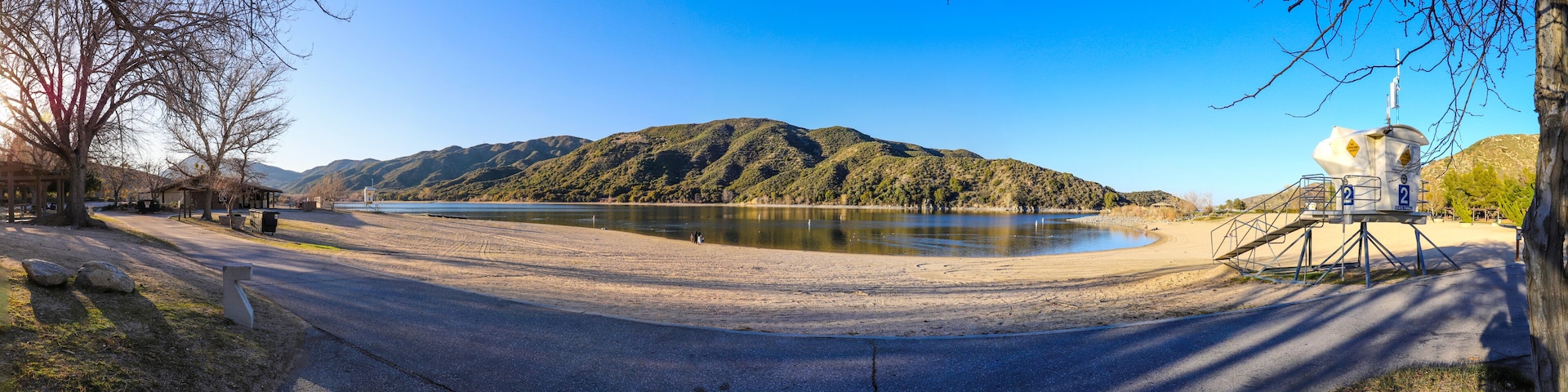 A lakeside road with a sandy beach, a forested hill, and a lifeguard tower.