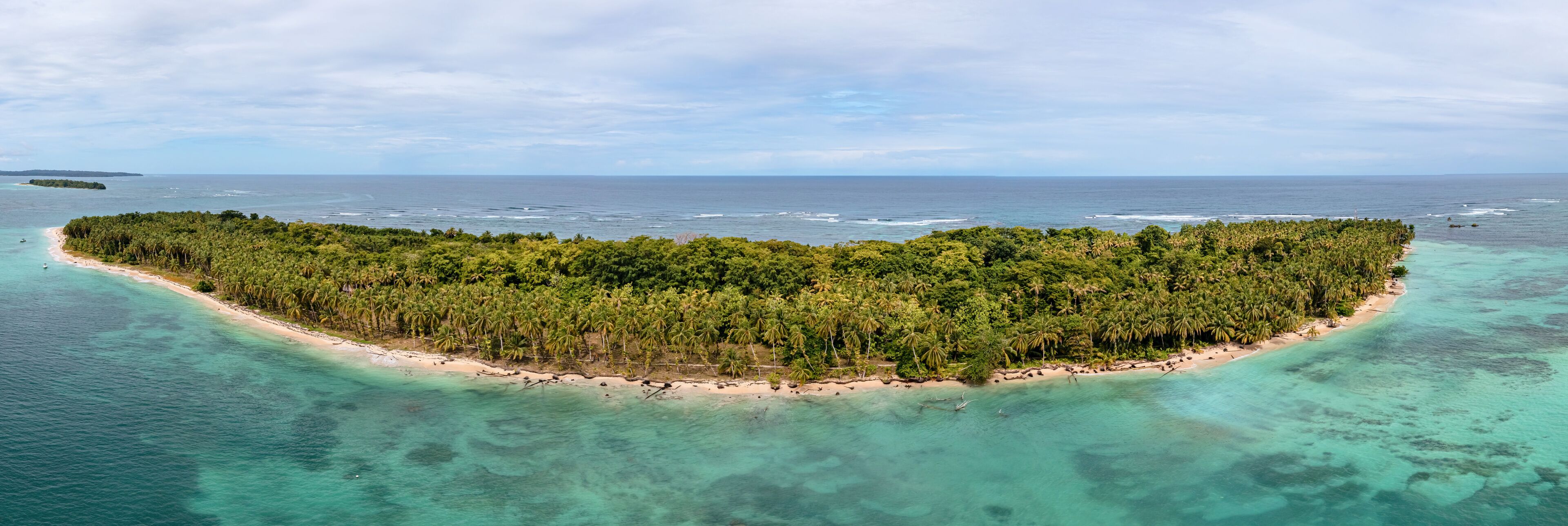 An aerial view of a tropical island with a narrow beach, dense palm trees, and clear turquoise waters.