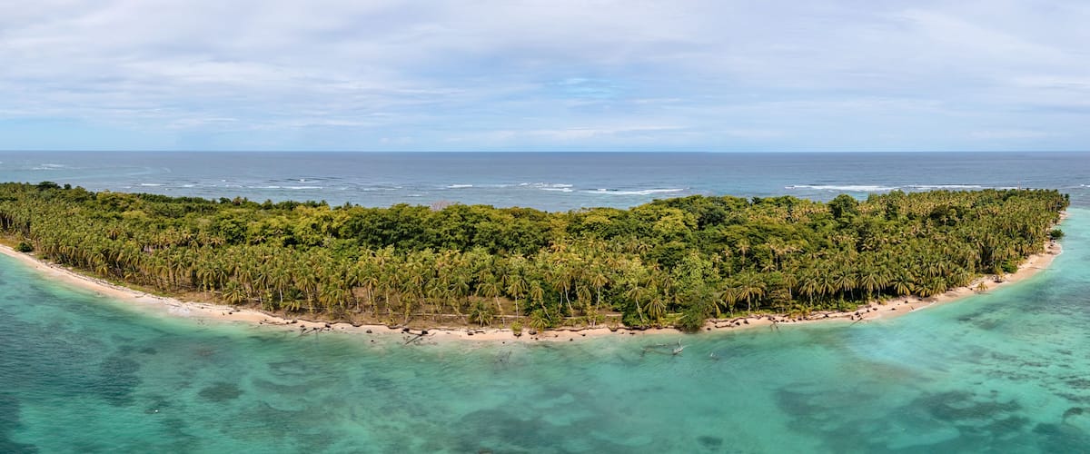 An aerial view of a tropical island with a narrow beach, dense palm trees, and clear turquoise waters.