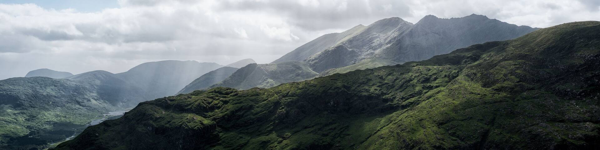 A scenic view showing the rolling green hillsides and imposing mountains of the Gap of Dunloe under a sky filled with dramatic clouds. Rain shafts are visible in the distance.