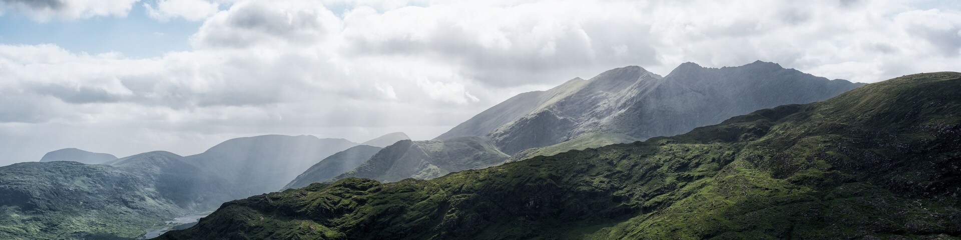 A scenic view showing the rolling green hillsides and imposing mountains of the Gap of Dunloe under a sky filled with dramatic clouds. Rain shafts are visible in the distance.