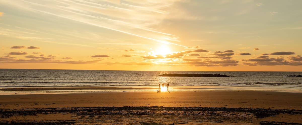 A beach at sunset with the sun low on the horizon, waves, and a rocky outcrop in the distance.