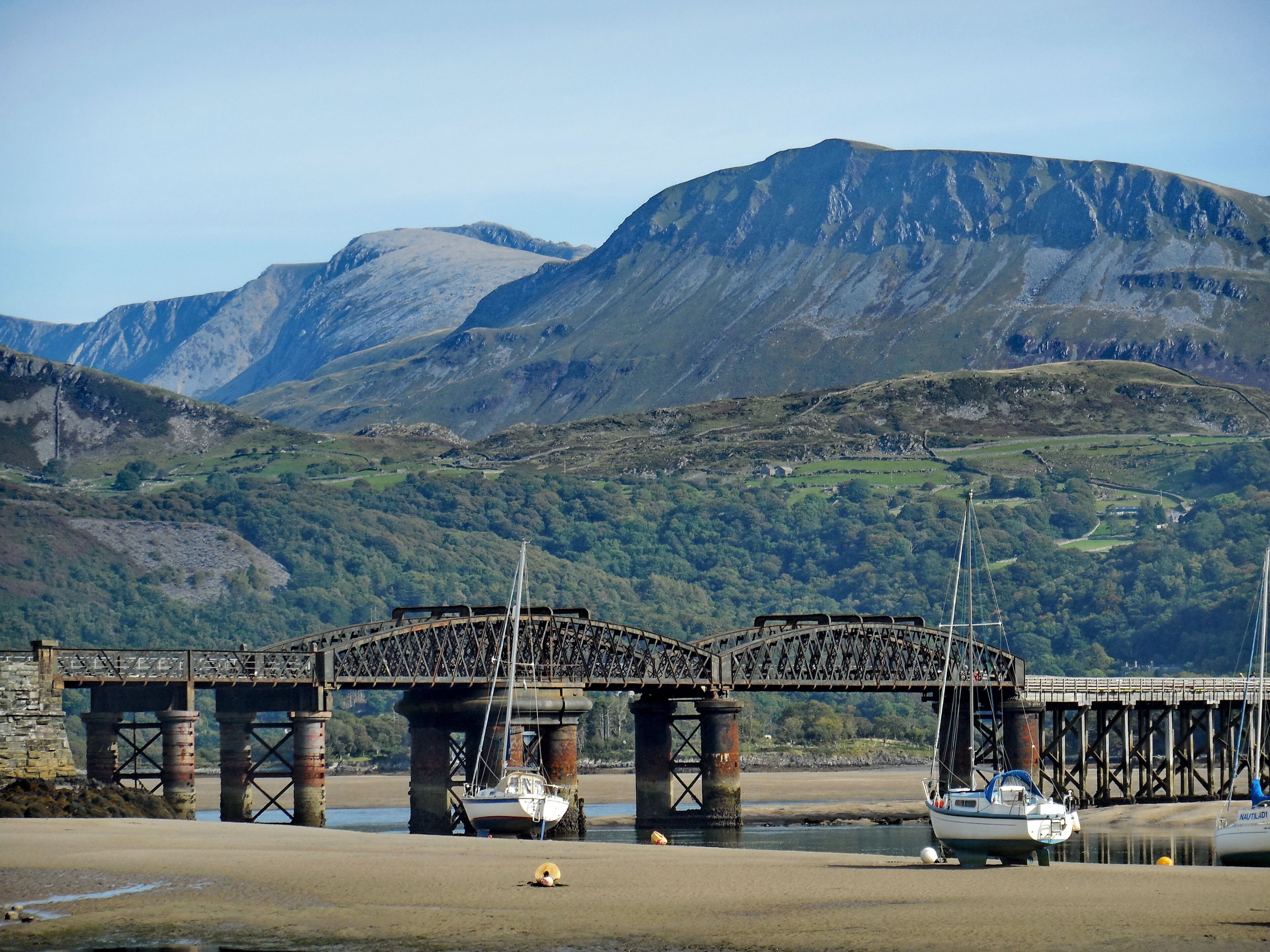 A bridge over a river with boats, a sandy shore, and mountains in the background.