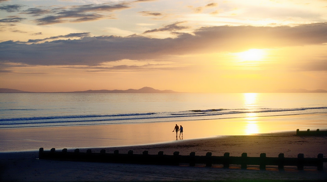 A beach at sunset with two people walking along the shore, waves gently crashing, and a distant mountain range.