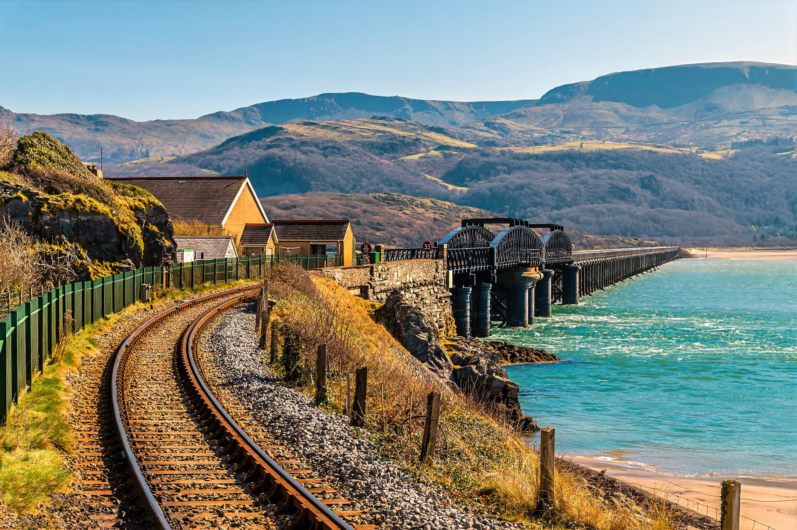 A coastal railway track with a bridge over water, surrounded by hills and a small building.